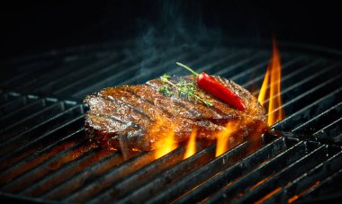 Hot spicy chili steak grilling on a summer barbecue over the flaming hot coals garnished with a whole red cayenne pepper and fresh rosemary clipart