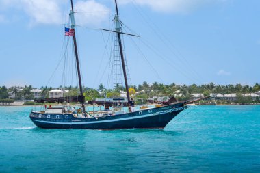 Key West, ABD - 04.30.2017: İnsanlar Meksika Körfezi 'nin turkuaz sularına yelken açan ahşap bir yatla denize açılıyorlar. Arka planda tropik tatil beldeleri ve ağaçlar var. Tropik cennette tatil. Karayip atmosferi.