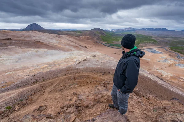 A man overlooking volcanic Icelandic landscape from the top of Namafjall. Dramatic clouds in the sky. Road in the distance winds through the land. Hverir, boiling mud area.