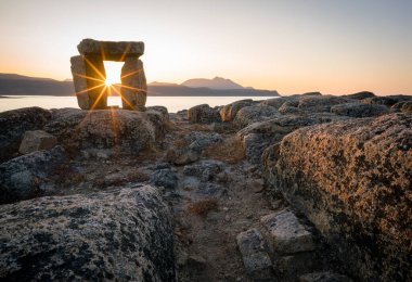 Sunset through Inuksuk in rough arctic landscape. Sunstar on the horizon in the fjord of Qikiqtarjuaq, Broughton Island, Nunavut. The north. clipart