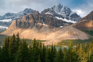 Kanada Banff Ulusal Parkı 'ndaki Bow Gölü' nün yukarısındaki Akbaba Tepesi kuleleri dramatik bulutların altında birkaç ışık huzmesi tarafından aydınlatılıyor. Kanada Kayalıkları 'nın göbeğinde bulutlu bir yaz günü. Tepesi karla kaplı uzun bir tepe..