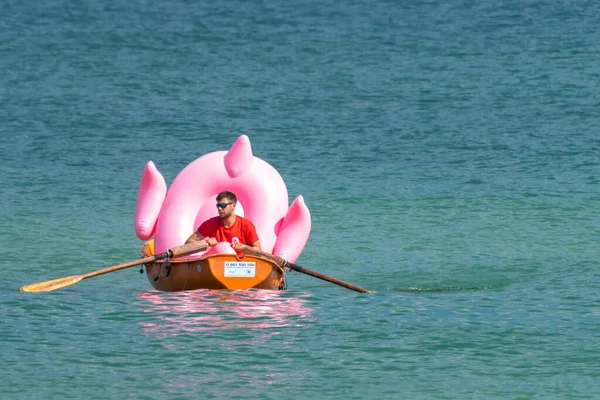 Hel, Polen - 08.01.2021: Bademeister rudert an einem sonnigen Sommertag in seinem orangefarbenen Boot und trägt einen großen rosa aufblasbaren Flamingo ans Ufer zurück. — Stockfoto