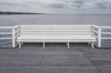 Gri bulutlu bir günde ahşap bir okyanus rıhtımına inşa edilmiş beyaz ahşap bank. Jurata Rıhtımı, Polonya, Baltık Sahili.