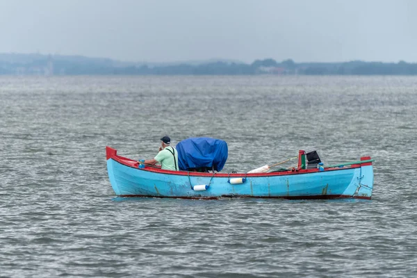 El pescador con camiseta verde fumando un cigarrillo se sienta frente a su pequeño barco azul, navegando hacia el mar Báltico cerca de Jastarnia, Polonia, en un día nublado de verano. La tierra en la neblina en el horizonte lejano. — Foto de Stock