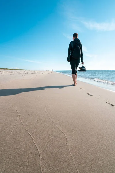 Foto vertical de una mujer en ropa oscura al aire libre con zapatos en la mano derecha, caminando descalza lejos del espectador en una playa. Hermosa mañana de verano junto al mar Báltico. Paseo costero de verano. — Foto de Stock