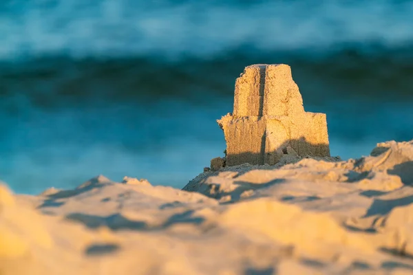 Una pequeña torre de castillo de arena construida en una playa con marea azul en el fondo borroso en la madrugada de verano. Diversión en la costa báltica en Hel, Polonia. Hora dorada en la playa. — Foto de Stock