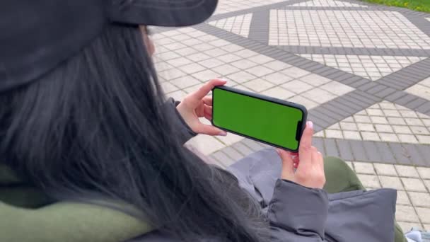 女の子ですキャップ保持電話で緑の画面がベンチに座って. — ストック動画