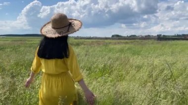 Hasır şapkalı ve uzun elbiseli bir kız yeşil bir çayırda yürüyor..