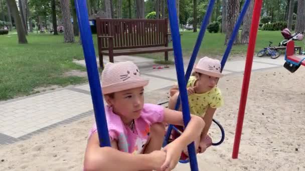 4k Sorelle gemelle tristi che dondolano su un'altalena in un parco giochi. — Video Stock