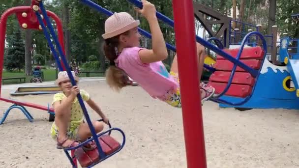 4k Sorridenti gemelle che dondolano su un'altalena in un parco giochi. — Video Stock