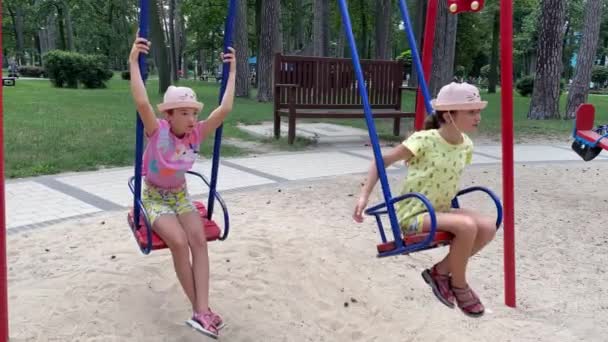 4k Hermanas gemelas sonrientes balanceándose en un columpio en un patio de recreo. — Vídeo de stock