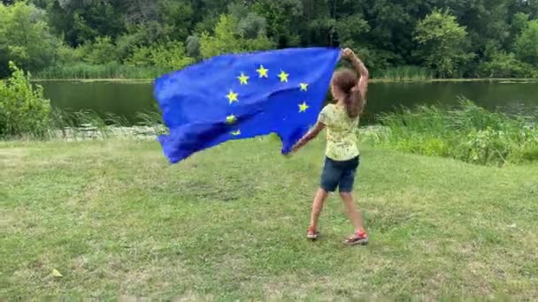 4k Petite fille souriante avec un grand drapeau de l'Union européenne tournant dans un parc. — Video