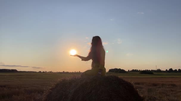 4k Silhueta de uma mulher sentada em um palheiro no campo e segurando o sol na mão. — Vídeo de Stock