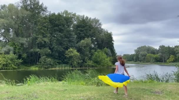 4k Μικρό κορίτσι στέκεται με μεγάλη ουκρανική σημαία κοντά στο ποτάμι στο πάρκο. — Αρχείο Βίντεο