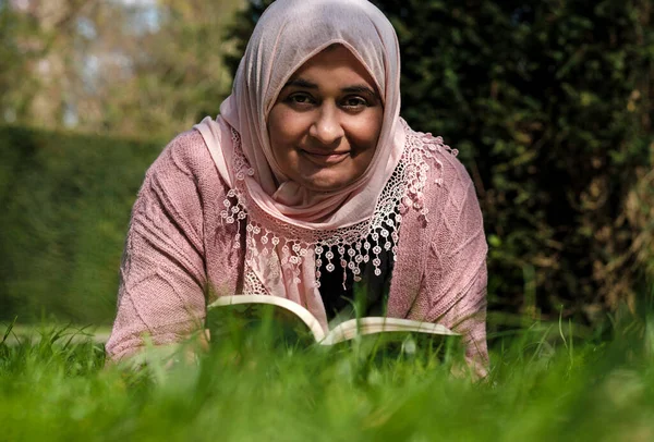 Mature muslim woman wearing hijab reading a book lying down on the grass. She is smiling.