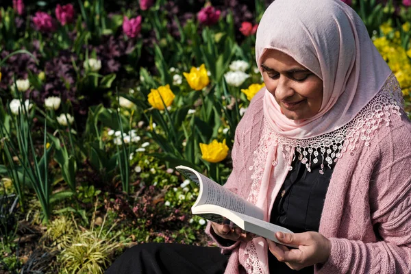 Mature muslim woman wearing hijab reading a book in a beautiful day in a park with flowers.