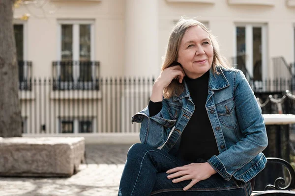 Kot pantolon giymiş olgun bir kadın dalgın görünüyor, bankta oturuyor. Arka planda bir ev.