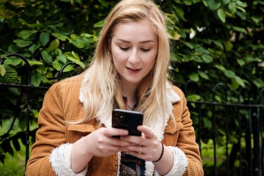 Akıllı telefonunu kullanan genç bir kadının portresi. Kahverengi bir ceket giyiyor ve ekrana vuruyor...