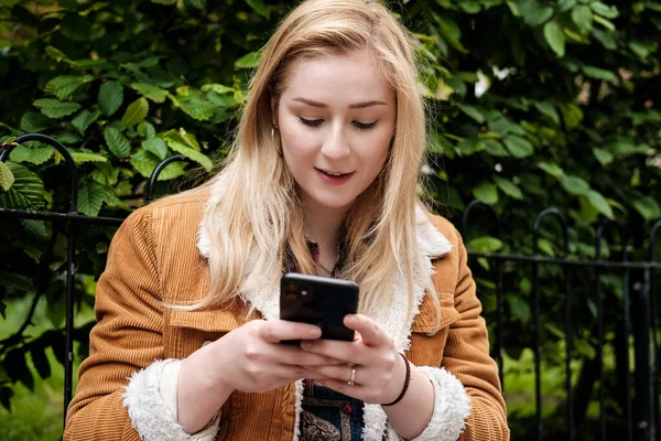 Akıllı telefonunu kullanan genç bir kadının portresi. Kahverengi bir ceket giyiyor ve ekrana vuruyor...