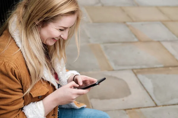 Genç sarışın kadın telefonunu kullanıyor ve gülüyor. Oturuyor ve eğleniyor..