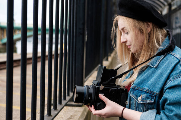 Young woman taking pictures with a medium format film camera. She is wearing a denim jacket and a black beret. Looking to camera.