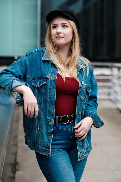 Köprü tırabzanına dayanmış genç bir kadının portresi. Kot ceket ve siyah şapka giyiyor..