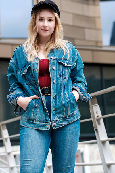 Portrait of young woman posing with a banister behind her. She is wearing denim jacket and trousers.