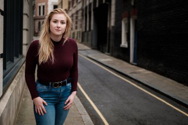 Dar bir sokakta poz veren genç bir kadının portresi. Üzerinde kestane rengi kazak ve kot pantolon var..