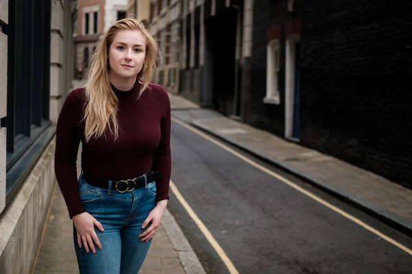 Dar bir sokakta poz veren genç bir kadının portresi. Üzerinde kestane rengi kazak ve kot pantolon var..