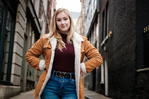 Dar bir sokakta kameraya bakan genç bir kadının portresi. Kestane rengi kazak, kadife ceket ve kot pantolon giyiyor. Sokaklar taş döşeli..