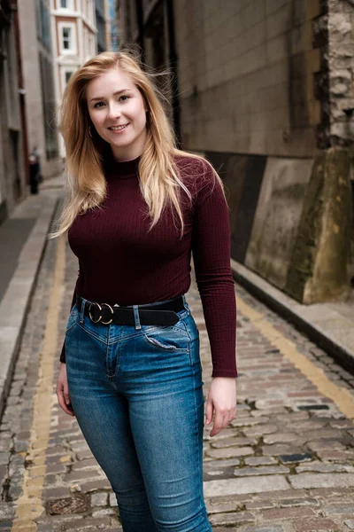 Çok dar bir sokakta poz veren gülümseyen genç bir kadın. Üzerinde kestane rengi kazak ve kot pantolon var. Sokaklar taş döşeli..