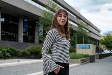 Arkasında bir binayla kameraya poz veren genç doğal bir kadın. Gülümsüyor ve çizgili kazak giyiyor..