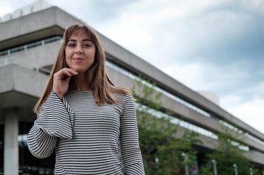 Arkasında bir binayla kameraya poz veren genç doğal bir kadın. Gülümsüyor ve çizgili kazak giyiyor..