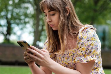 Çimlerin üzerinde telefonunu kullanan romantik genç bir kadının portresi. Bahar elbisesi giyiyor ve telefonuna bakıyor..