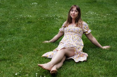 Bir bahar günü çimlerin üzerinde oturan rahat genç bir kadın. Bahar elbisesi giyiyor..