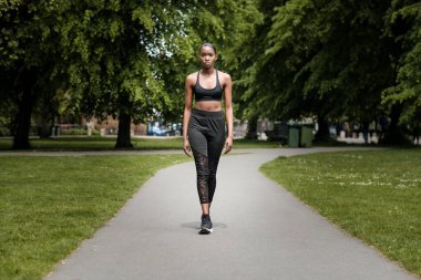 Siyah genç bir kadın parkta egzersiz yapıyor. Kameraya doğru emin adımlarla yürüyor..