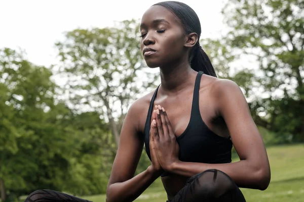 Genç siyahi kadın parkta meditasyon ve yoga yapıyor. Siyah spor kıyafeti giyiyor..