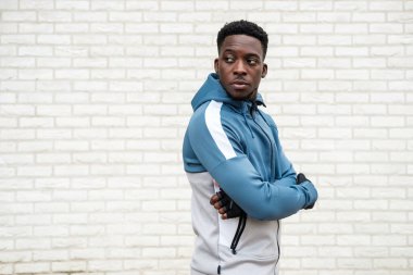 Beyaz tuğla bir duvarın önünde poz veren zinde bir zenci. Beyaz ve mavi bir kapüşonlu giyiyor..