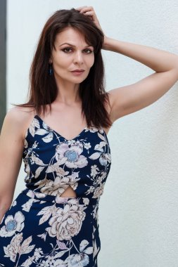 Çekici, olgun bir kadın beyaz bir duvara karşı poz veriyor. Bahar elbisesi giyiyor..