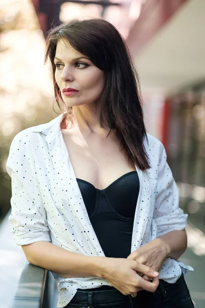 Çekici, olgun bir kadın şehir ortamında poz veriyor. Beyaz bir bluz ve siyah bir korse giyiyor..