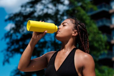 Sarı şişeden içen çekici genç bir kadın. Sıcak bir yaz gününde siyah bir bluz giyiyor. Karışık ırk.