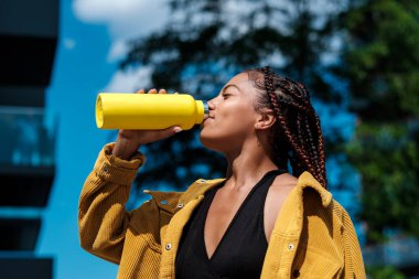 Sarı şişeden içen çekici genç bir kadın. Sarı bir ceket giyiyor ve bugün yazın sıcak bir gün. Gülümsüyor. Karışık ırk.