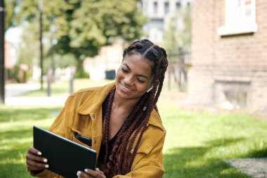 Parkta tablet kullanan havalı saçlı genç bir kadın. Çimlerin üzerinde oturuyor ve eğleniyor..