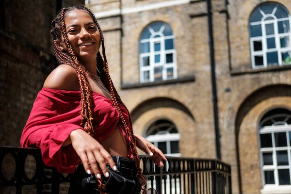 Sıcak bir yaz gününde Londra sokaklarında poz veren havalı saçlı genç bir kadın. Örgülü ve kırmızı bluz giyiyor..