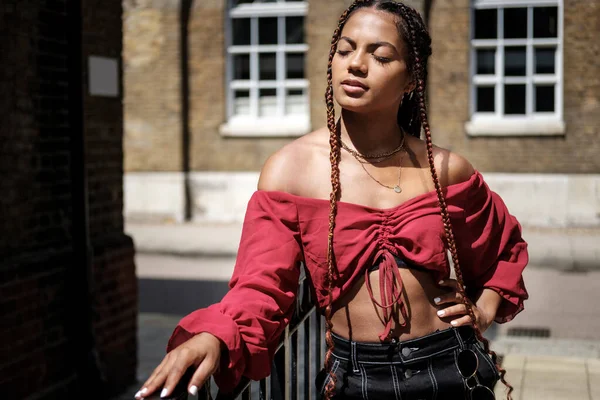 Sıcak bir yaz gününde Londra sokaklarında poz veren havalı saçlı genç bir kadın. Örgülü ve kırmızı bluz giyiyor..