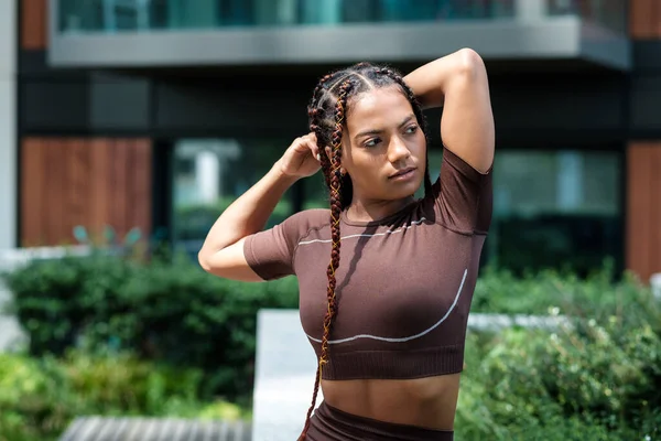 Spor kıyafetleri giyen ve şehir ortamında esneme hareketleri yapan genç bir kadın. Karışık ırk, örgü..