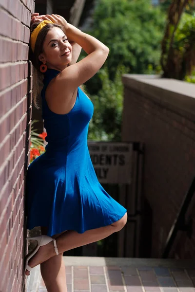 An attractive young woman is posing leaning against a brick wall. She is wearing a blue spring dress in a sunny summer day.