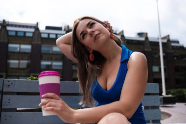 Çekici genç bir kadın bankta kahve içiyor. Mavi bir bahar elbisesi giyiyor ve elinde bir kupa tutuyor..