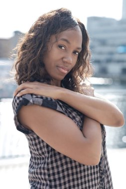 Olgun siyah, arkasında güneş varken poz veriyor. Kollarını kavuşturmuş ve kıvırcık saçları var..