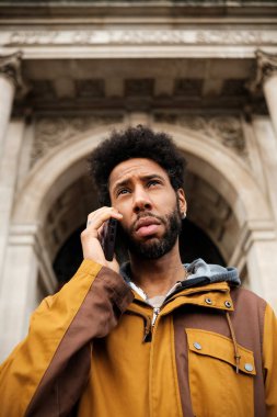 Afro saçlı genç bir adam, kolonları ve kemerleri olan, kahverengi ve oklu bir ceket giyen, anıtsal bir binanın önünde telefon görüşmesi yapıyor.